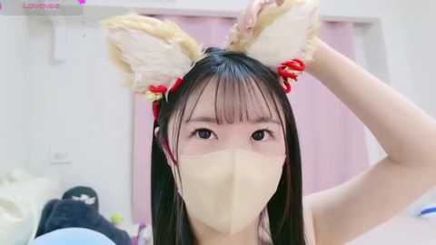 Media: Video of an East Asian woman wearing a beige face mask, bunny ears, and red ribbons, indoors with white walls, pink curtains, and a bed.