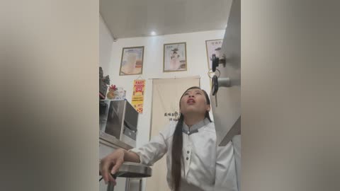 Video of a young Asian man in a white chef's uniform, standing in a small, tidy kitchen with white walls, framed posters, and a microwave on a counter.