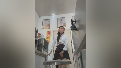 Media: Video of a petite Asian woman with long black hair in a white blouse and black skirt, standing in a small, dimly lit kitchen with a metal stool.