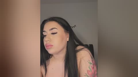 Media: Video of a Latina woman with long black hair, wearing pink lipstick and black eyeliner, leaning against a beige wall. She has a tattoo of red roses on her right arm.