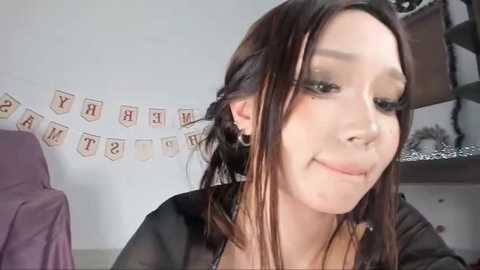 Media: A video of a young woman with fair skin, brown hair, and subtle makeup, wearing a black top, smiling slightly. Background includes a white wall with a decorative banner, a purple chair, and a cluttered shelf.