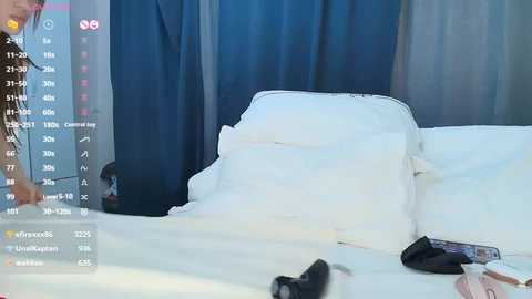 Media: Video of a bed with white sheets and pillows, featuring a person partially visible in the background. The room has dark blue curtains and a black object on the floor.