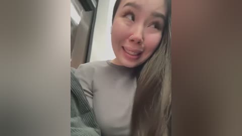 Media: A video of an Asian woman with long brown hair, wearing a light grey sweater, smiling slightly, taken in a narrow, dimly-lit corridor.