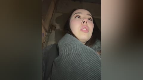 Media: A video of a young woman with long, straight brown hair, wearing a dark green, ribbed sweater, looking upward with a slight pout. The background is dimly lit, showing a textured, possibly concrete wall.