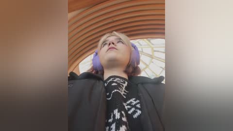 Media: Video of a young woman with light skin and blonde hair, wearing purple headphones, a black jacket, and a black sweater with white snowflake patterns, standing under a wooden arched ceiling in a modern, light-filled indoor space.