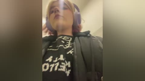 Media: A low-angle video of a woman with short, blonde hair, wearing purple headphones and a black graphic tee with white text. She has a confident expression and is indoors.