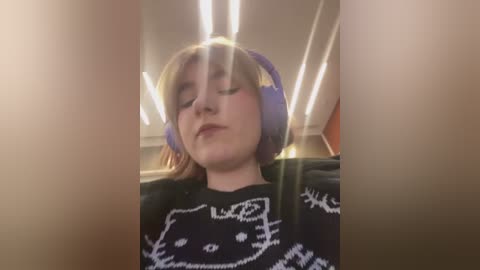 Media: A close-up video of a young woman with fair skin, short blonde hair, and closed eyes, wearing purple over-ear headphones and a black sweater with a white cat design. The background features a ceiling with fluorescent lights.