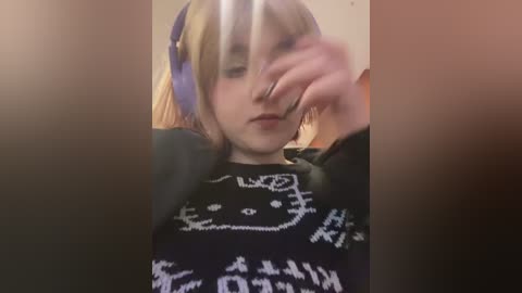 Media: Video of a young woman with blonde hair, wearing a black sweater with a white cat design and purple headphones. She covers her face with her hand, looking slightly to the side.