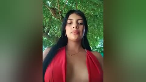 Media: Video of a young, dark-skinned woman with long black hair, wearing a revealing red halter top, standing in a lush green forest.