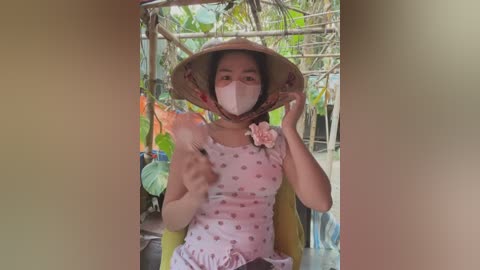 Media: Video of a woman with light skin and dark hair wearing a conical hat, pink floral dress, and face mask, seated on a wooden bench under a bamboo roof, surrounded by greenery.