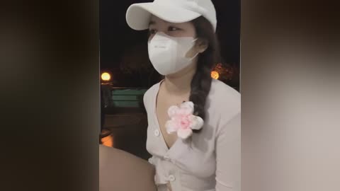 Media: Video of an Asian woman with light skin and long, dark hair in a braid, wearing a white cap, mask, and floral headband, standing outdoors at night with blurred background lights.