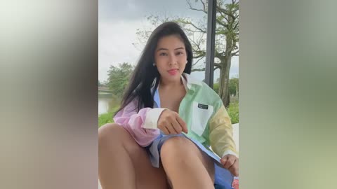 Media: Video of a young Asian woman with long black hair, wearing a colorful, oversized jacket, sitting on a balcony with a serene outdoor view in the background.