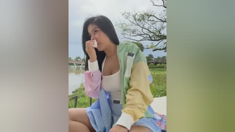 Media: Video of a young woman with long black hair, wearing a pastel-colored jacket over a white top, sitting outdoors near a pond, holding a tissue to her mouth, with a blurred background.