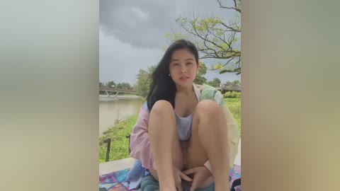 Media: A video of an Asian woman with long black hair, wearing a light blue top and pastel-colored jacket, sitting on a colorful blanket by a serene lake with a cloudy sky and lush greenery in the background.