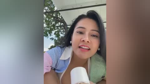 Media: Video of a smiling Asian woman with straight black hair, wearing a colorful striped top, holding a white cup, surrounded by greenery and modern architecture.