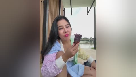 Media: Video of a young Asian woman with long black hair, wearing a colorful striped shirt, sitting outdoors, eating a chocolate bar. Background shows a modern building and water.