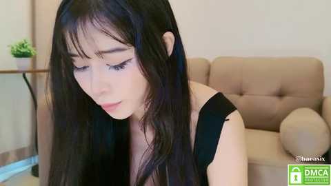 Media: Video of a young Asian woman with long black hair and fair skin, wearing a black top, sitting on a beige couch in a modern, indoor setting.