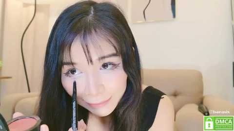 Media: Video of a young Asian woman with long black hair, applying eyeliner with a black brush, in a light-colored room with minimalistic decor.