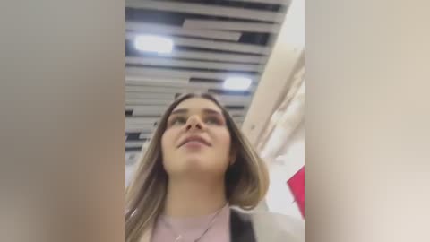 Media: A video of a young woman with long, light brown hair, wearing a pink top, standing in a modern, brightly lit indoor space with white and black ceiling panels and red accents.