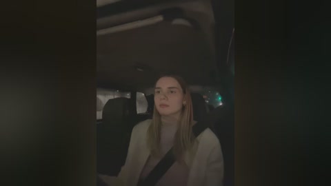 Media: Video of a young woman with long blonde hair, wearing a beige jacket, driving a car at night. The dimly lit interior features dark seats and a visible seatbelt.