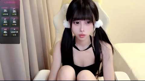 Media: A video of an Asian woman with long black hair in pigtails, wearing a black, low-cut top, sitting on a beige chair, with a virtual health app display in the background.