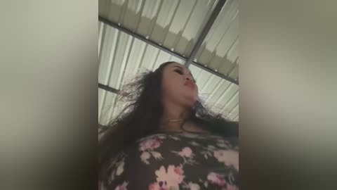 Media: Video of a woman with long, curly black hair, wearing a black floral dress, standing under a corrugated metal roof. Her facial expression is relaxed, with slightly parted lips and closed eyes.