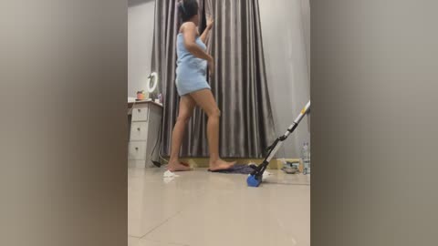 Media: Video of a woman in a light blue nightgown, barefoot, mopping the floor in a dimly lit, cluttered room with beige tiles, a white dresser, and a wall-mounted mirror.