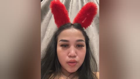 Media: Video of a young woman with long black hair, wearing red fluffy bunny ears, and a neutral expression. She is indoors, with a beige curtain in the background.