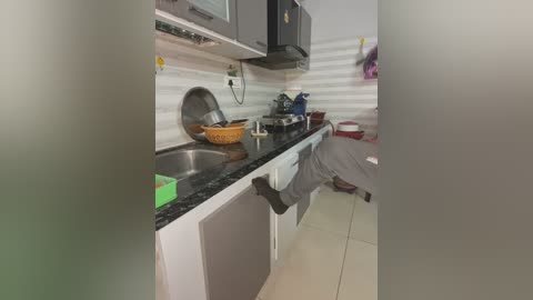 Media: A video of a small, cluttered kitchen with white cabinets, black countertops, and a person wearing gray pants leaning over the sink. The background shows a white-tiled backsplash and a microwave above the stove.