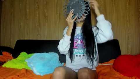 Media: A video of a young woman with long black hair wearing a gray sweatshirt, sitting on a bed with orange sheets. She holds a decorative, gray, circular mirror over her face, creating a playful, mysterious effect.