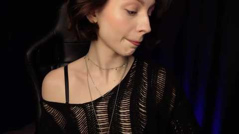 Media: Video of a fair-skinned woman with dark, wavy hair, wearing a black, off-shoulder knit sweater and layered gold necklaces, seated in a dimly lit, black background.