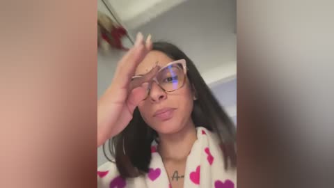 Video of a young woman with medium skin tone, long dark hair, wearing large pink-tinted glasses, and a white robe with pink heart patterns. She is adjusting her glasses, indoors with blurred background.