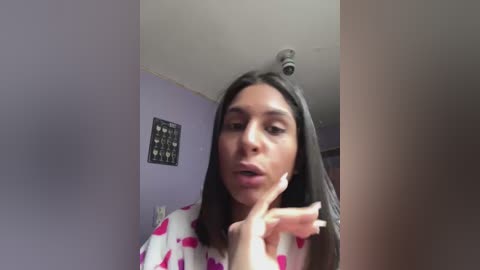 Media: Video of a Latina woman with medium brown skin, straight black hair, and wearing a white robe with pink elephants, making a dramatic pose with her hand near her face in a dimly lit room with purple walls and a framed heart collage.