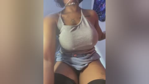 Media: Video of a curvy Black woman in a gray tank top and black shorts, sitting indoors with a blurred background.