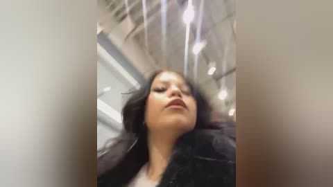 Media: Video of a woman with long black hair, wearing a dark jacket, looking upward in a dimly lit, modern indoor space with visible ceiling panels and recessed lighting.