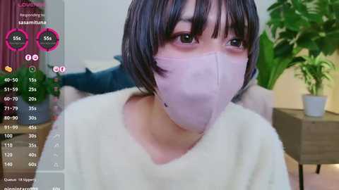 Media: A video of an Asian woman with short black hair, wearing a white mask, light pink shirt, and a green potted plant in the background.