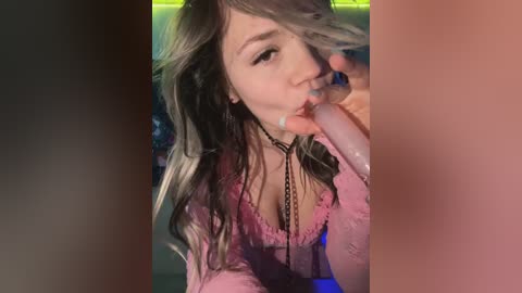 Media: A close-up video of a young woman with long, wavy hair, wearing a pink blouse, performing oral sex on a large, erect penis. The background is dimly lit with green and red lights.