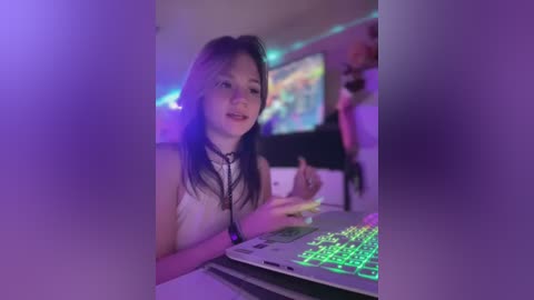 Media: Video of a young Asian woman with long black hair, wearing a white sleeveless top, typing on a colorful glowing keyboard in a dimly lit room with purple LED lights.