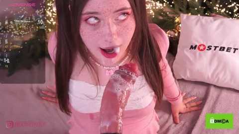 A video featuring a fair-skinned woman with long brown hair, wearing a pink sweater, sitting on a couch. She has a shocked expression with her mouth open, and a clear liquid is dripping from her chest. Background shows a festive room with Christmas decorations and a \"MOSTBEAUTIFUL\" pillow.