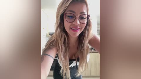 Media: A video of a smiling woman with long blonde hair, wearing glasses and a black-and-white striped top, leaning forward in a modern kitchen with white cabinets.