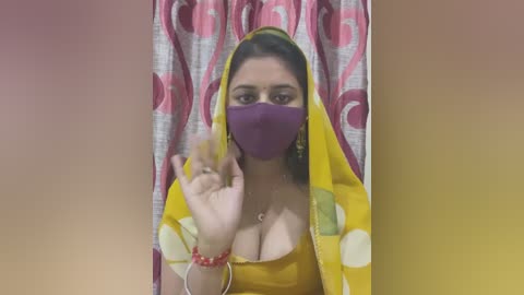 Media: Video of a South Asian woman with medium skin tone, wearing a yellow sari with purple mask and polka dot pattern, raising her hand. Background features a pink and white curtain.