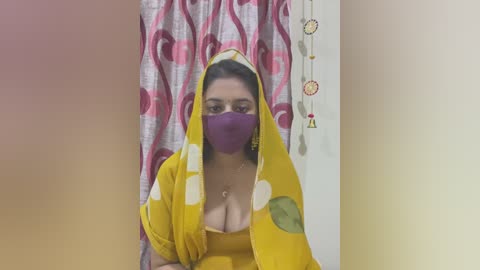 Media: Video of a woman with fair skin, wearing a yellow sari with white polka dots, purple face mask, and a deep neckline, in a room with pink and white patterned curtains and dreamcatcher decorations.