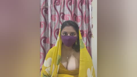 Media: Video of a South Asian woman with medium skin tone and dark hair, wearing a yellow saree, purple face mask, and green earrings, standing in front of a pink and white patterned curtain.