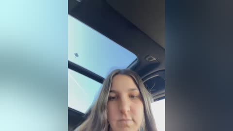 Media: Video of a young woman with long, straight brown hair, light skin, and a neutral expression, taken from inside a car. The background shows a bright, clear sky through the windshield.