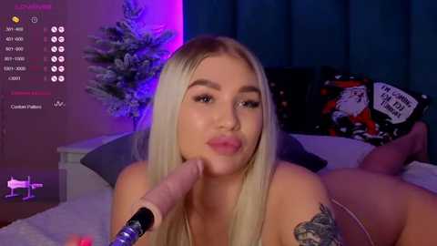 Media: Video of a blonde woman with light skin, wearing lingerie, lying on a bed. She has a tattoo on her arm and is performing oral sex on a dildo. Background includes a Christmas tree and festive decorations.