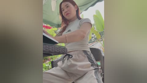 Media: Video of a young Asian woman with straight, shoulder-length red hair, wearing a grey crop top and white pants, sitting at an outdoor table under a green canopy.