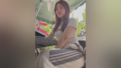 Media: Video of an Asian woman with long brown hair, sitting on a patio chair, wearing a beige t-shirt and striped pants, surrounded by lush green plants and a red flower pot.