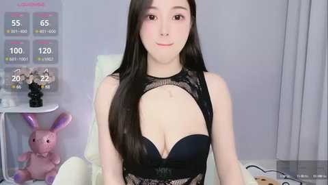 Media: A video of an East Asian woman with long black hair, wearing black lace lingerie, sitting in a white chair. The background features a gray wall, a digital calendar, and a plush bunny.