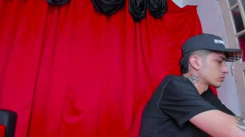 Media: Video of a tattooed man in a black cap and shirt, standing against a vibrant red curtain backdrop with black drapes.