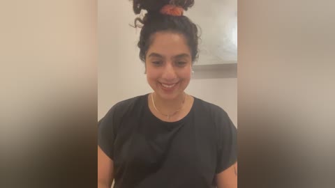 Media: Video of a South Asian woman with medium skin tone, wearing a black T-shirt, smiling, with her hair tied up in a bun. Background is a plain beige wall.